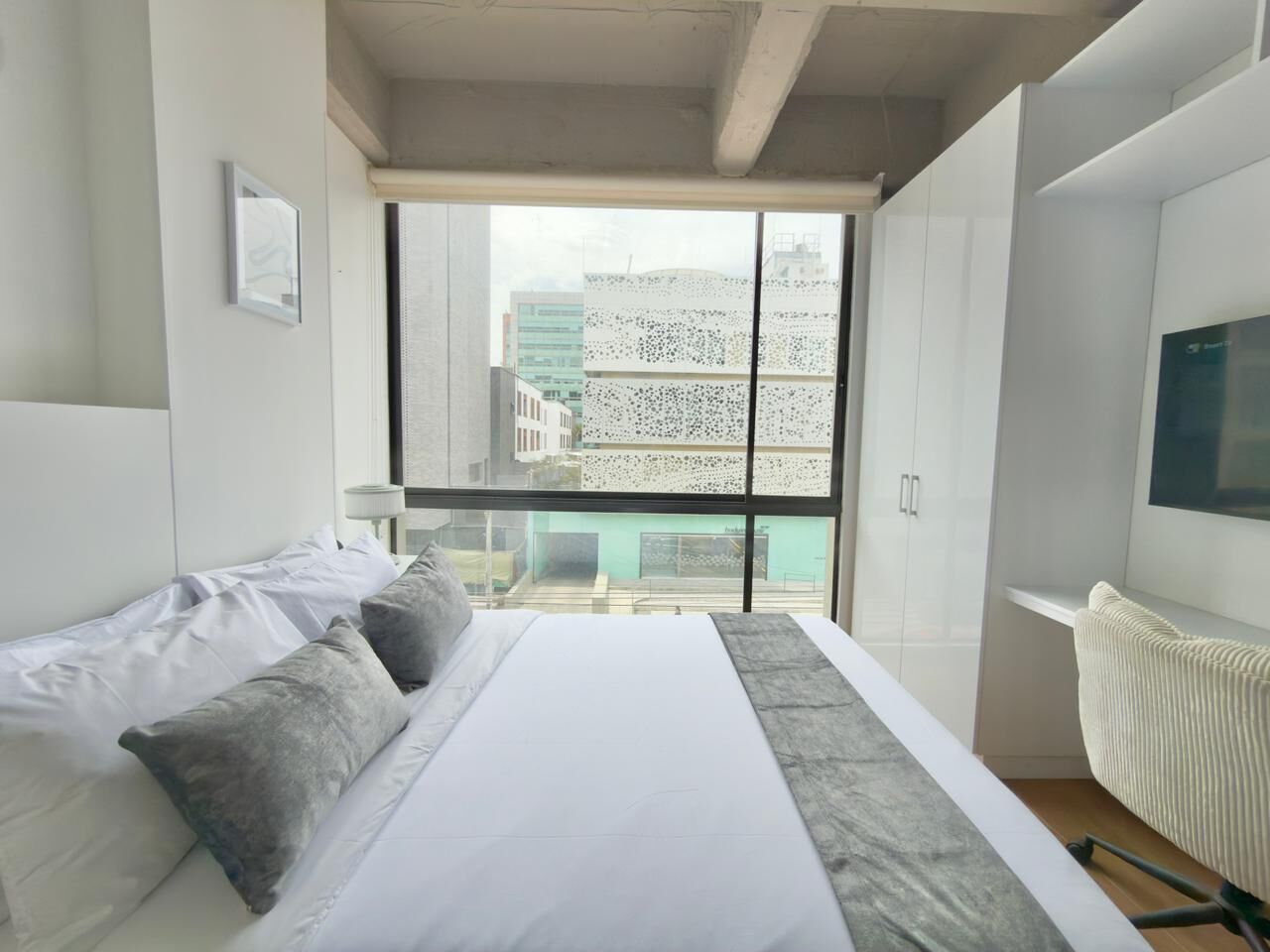 A modern hotel room with a large bed, a desk, and a view of buildings through a large window.