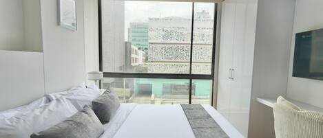 A modern hotel room with a large bed, a desk, and a view of buildings through a large window.