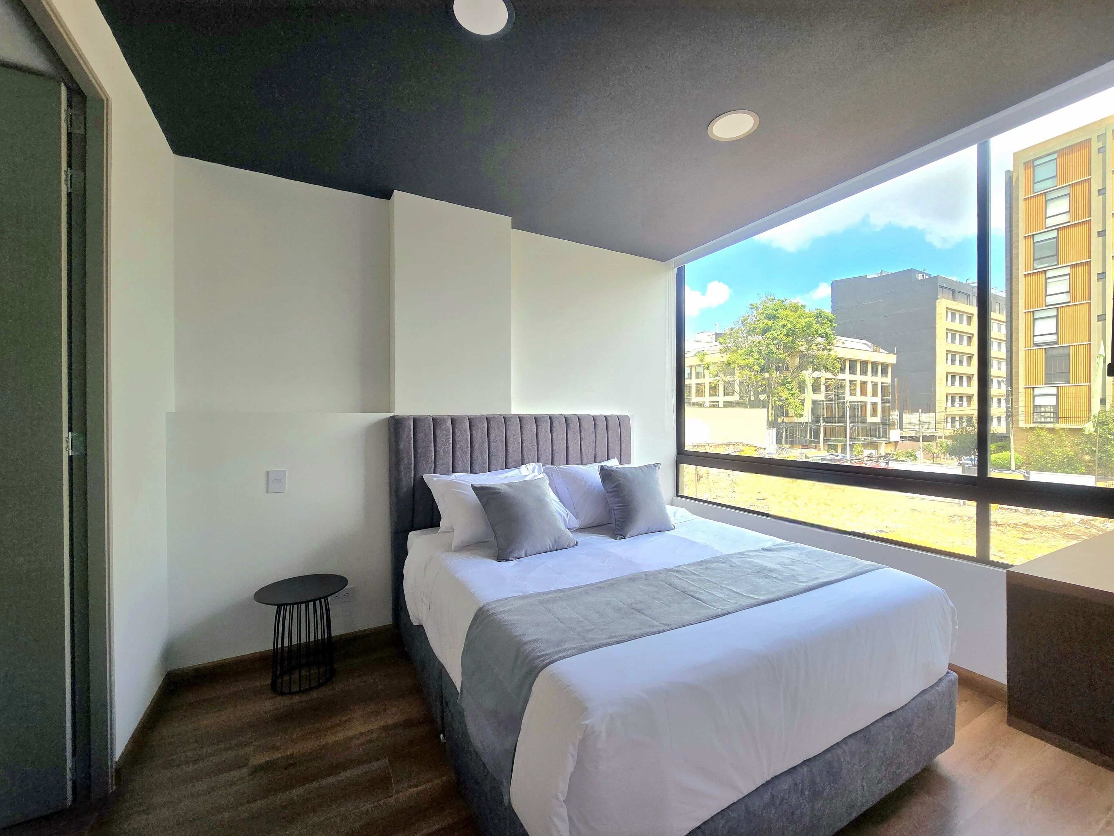 A modern hotel room with a large bed, a small side table, and a view of the cityscape through a large window.