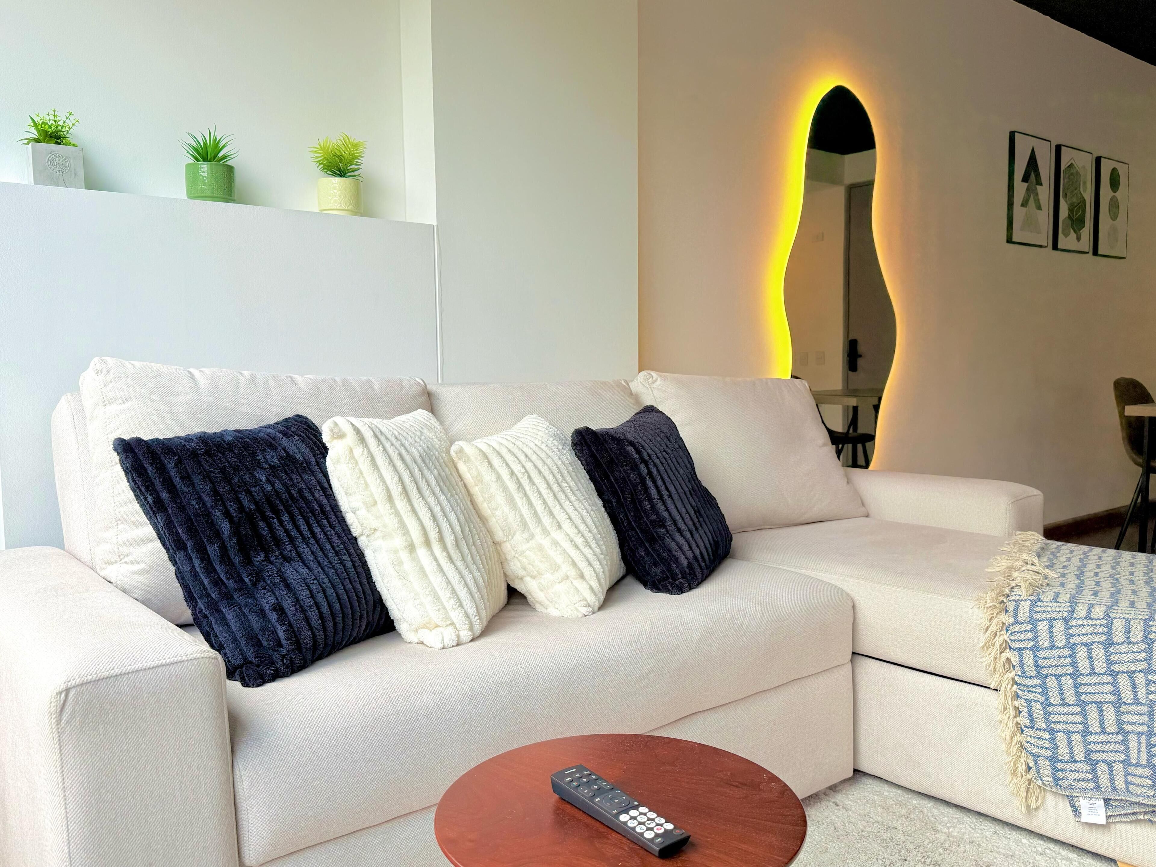 Een moderne woonkamer met een bank, een afstandsbediening op een salontafel en decoratieve planten op een plank.