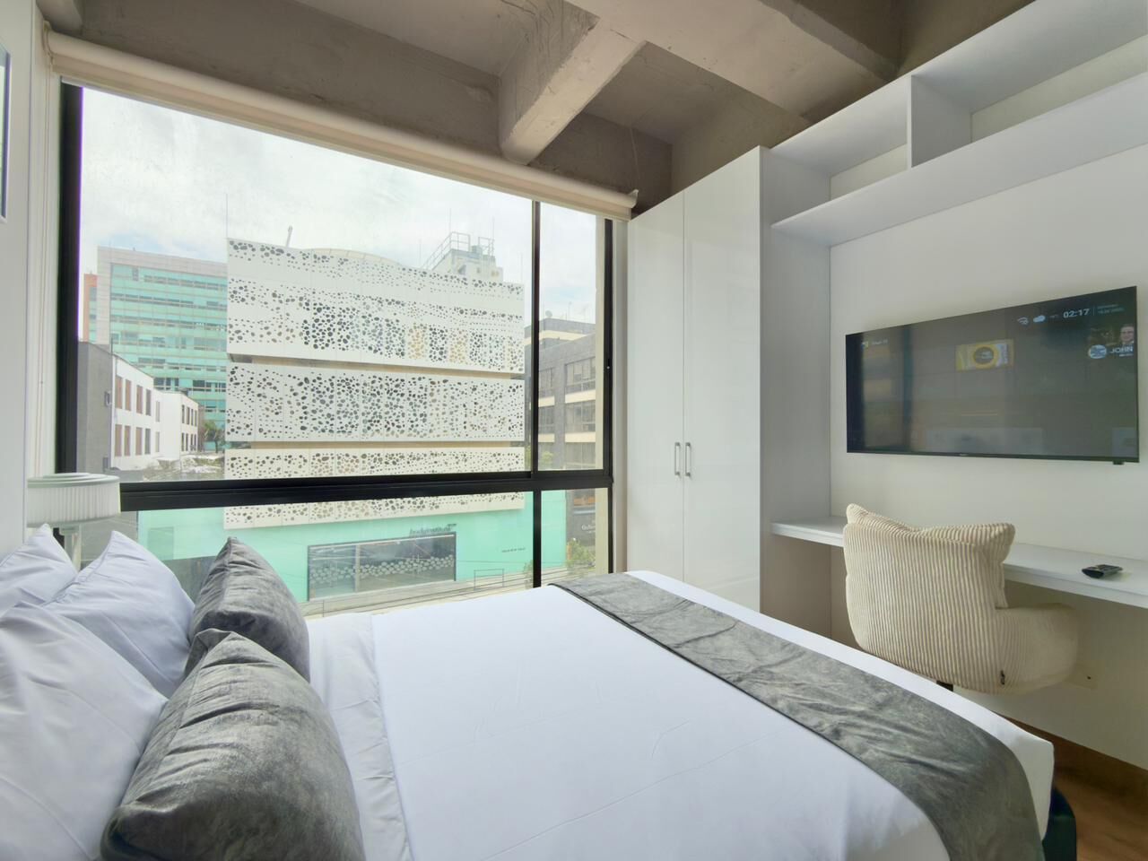 A modern hotel room with a large bed, a TV mounted on the wall, and a view of the cityscape through the window.