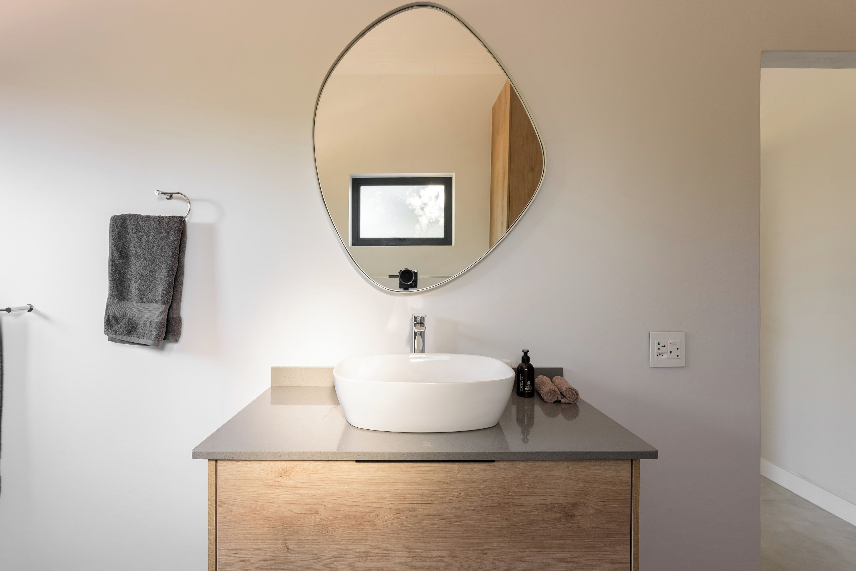 Ein Badezimmer mit rundem Spiegel, Waschbecken in Weiß, Holzschrank, Handtuchhalter mit Handtuch und einer kleinen Flasche.