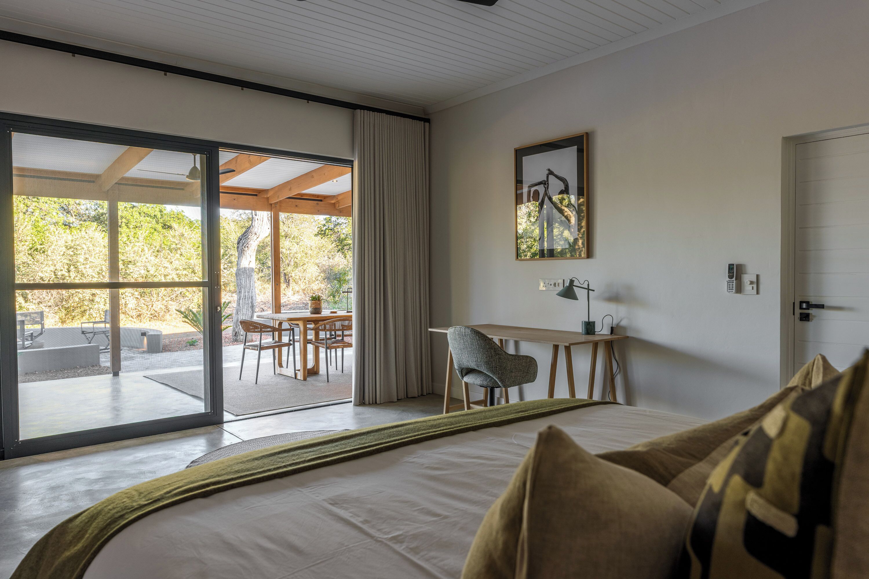A bedroom with a large bed, a desk, and a view through a sliding glass door to an outdoor patio.
