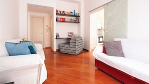 Un salon moderne avec un canapé blanc, un canapé rouge et un pouf à motifs. On y trouve une bibliothèque remplie de livres et d’objets décoratifs.