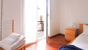 Une chambre d’hôtel avec un lit, une table de chevet avec une lampe, une commode en bois, une chaise avec une serviette, et une vue sur l’extérieur par une porte ouverte.