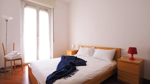 Un lit bien fait, avec des draps blancs, un plaid bleu et une lampe de chevet rouge.