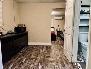 A compact room with a wooden floor, a bed with a red bedspread, a small desk with a lamp, a microwave, and a wall-mounted air conditioner. - Unit 2 - Private Queen Studio Near Miami Airport & Wynwood (Miami)
