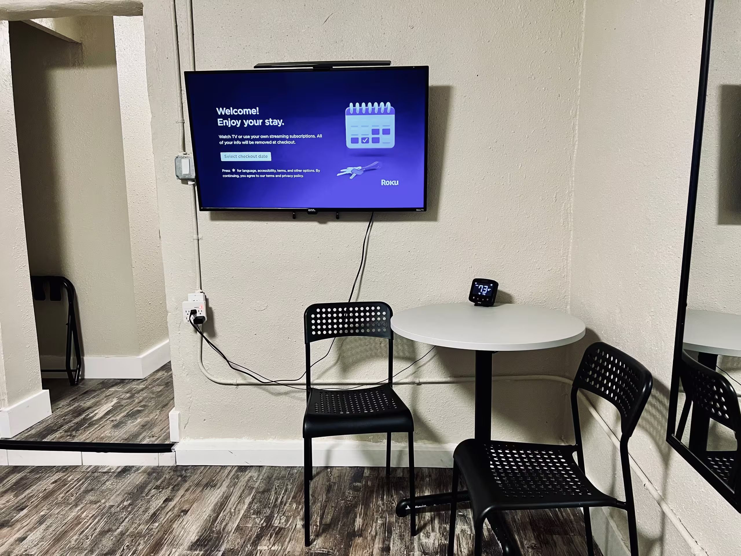 A room with a flat-screen TV mounted on the wall displaying a welcome message, a small table with a digital clock, and two black chairs.