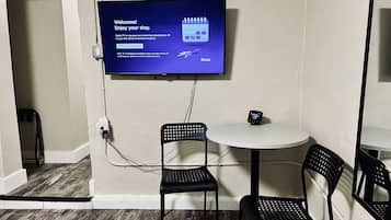 A room with a flat-screen TV mounted on the wall displaying a welcome message, a small table with a digital clock, and two black chairs.
