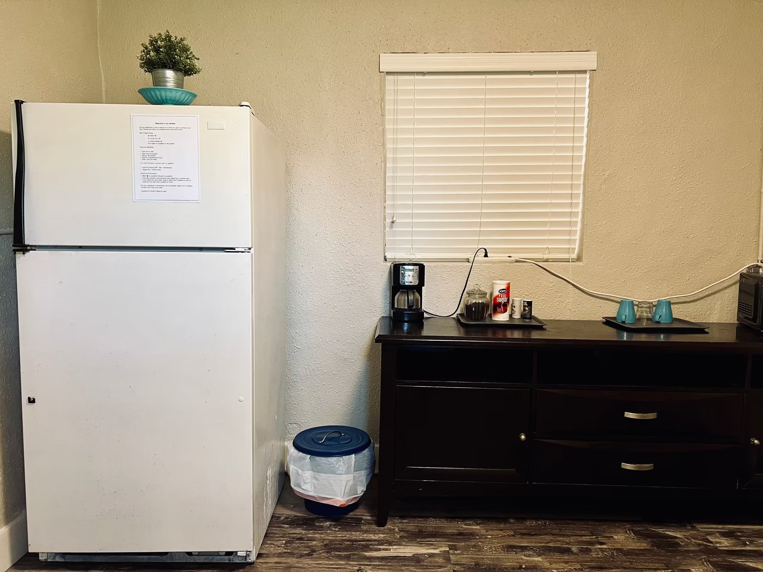 A compact kitchenette with a white refrigerator, a coffee maker, a can of sweetener, and a trash bin.