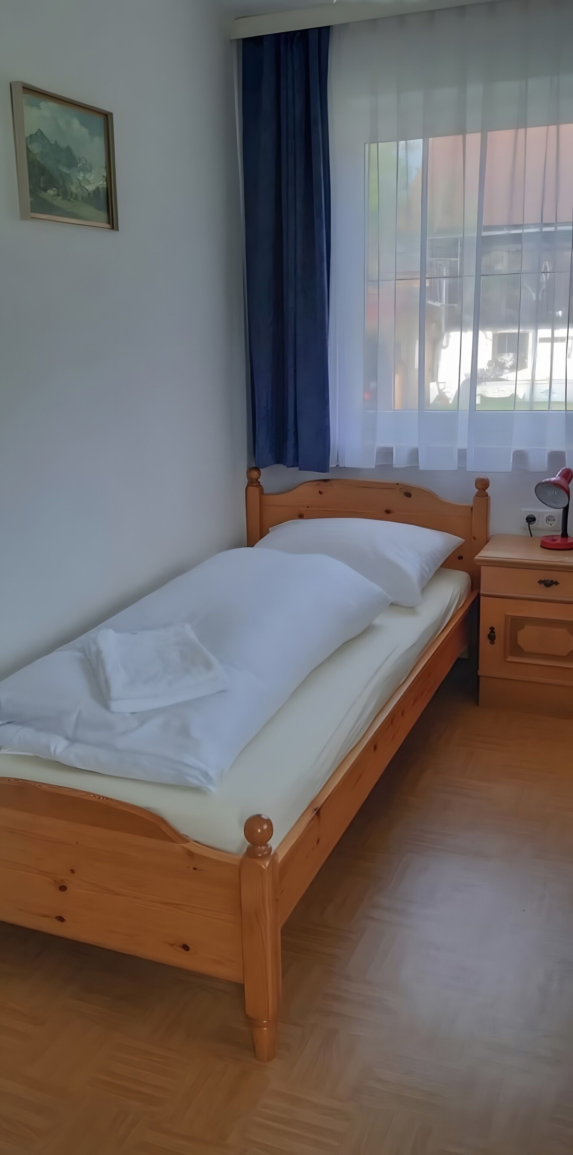 Een enkel bed met houten frame, een nachtkastje met lamp en een raam met gordijnen.