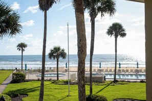 A pool area with palm trees, a view of the ocean, and a fenced deck with lounge chairs. - Renovated Beach Condo Balcony, Pool/BBQ | CoqB202 (St. Augustine)