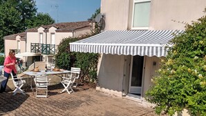 A patio with a table and chairs, a striped awning, and a building with a door and window. - House in lovely gardens with shared pool in Dordogne, parking on site (Bouteilles Saint-Sébastien)