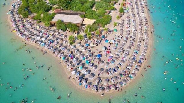 An aerial view of a beach with numerous umbrellas and people, surrounded by clear turquoise waters and green hills. - NG Hisar Apart (Fethiye)