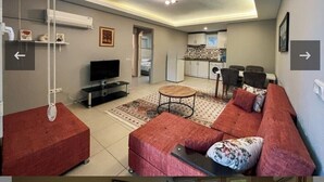 A modern living room with a sofa, a coffee table, and a television. There is a kitchen area in the background with cabinets and a countertop. - NG Hisar Apart (Fethiye)
