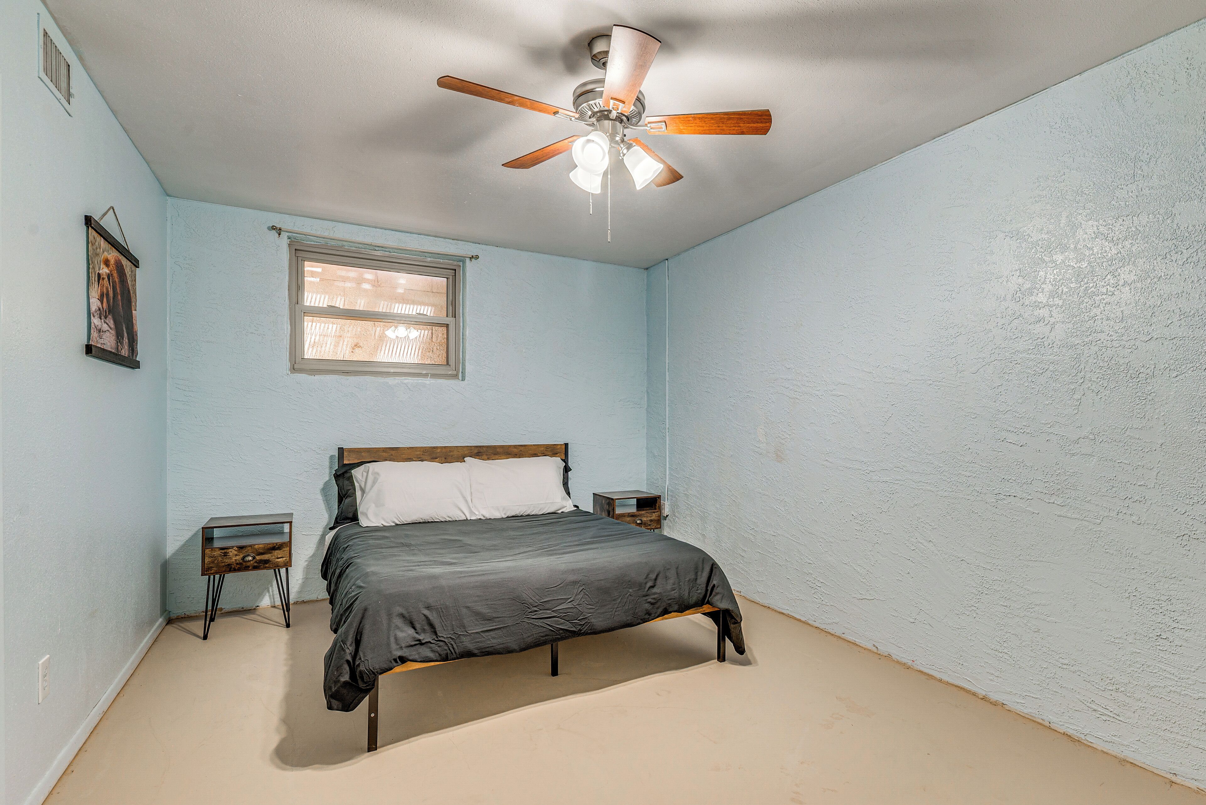 Et soveværelse med seng, ventilator, natbord, vindue og et billede på væggen.