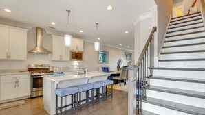 A modern kitchen with white cabinetry, a central island with bar stools, stainless steel appliances, and a staircase leading to another level. - Walk to Ocean: Group Getaway in Virginia Beach! (Virginia Beach)