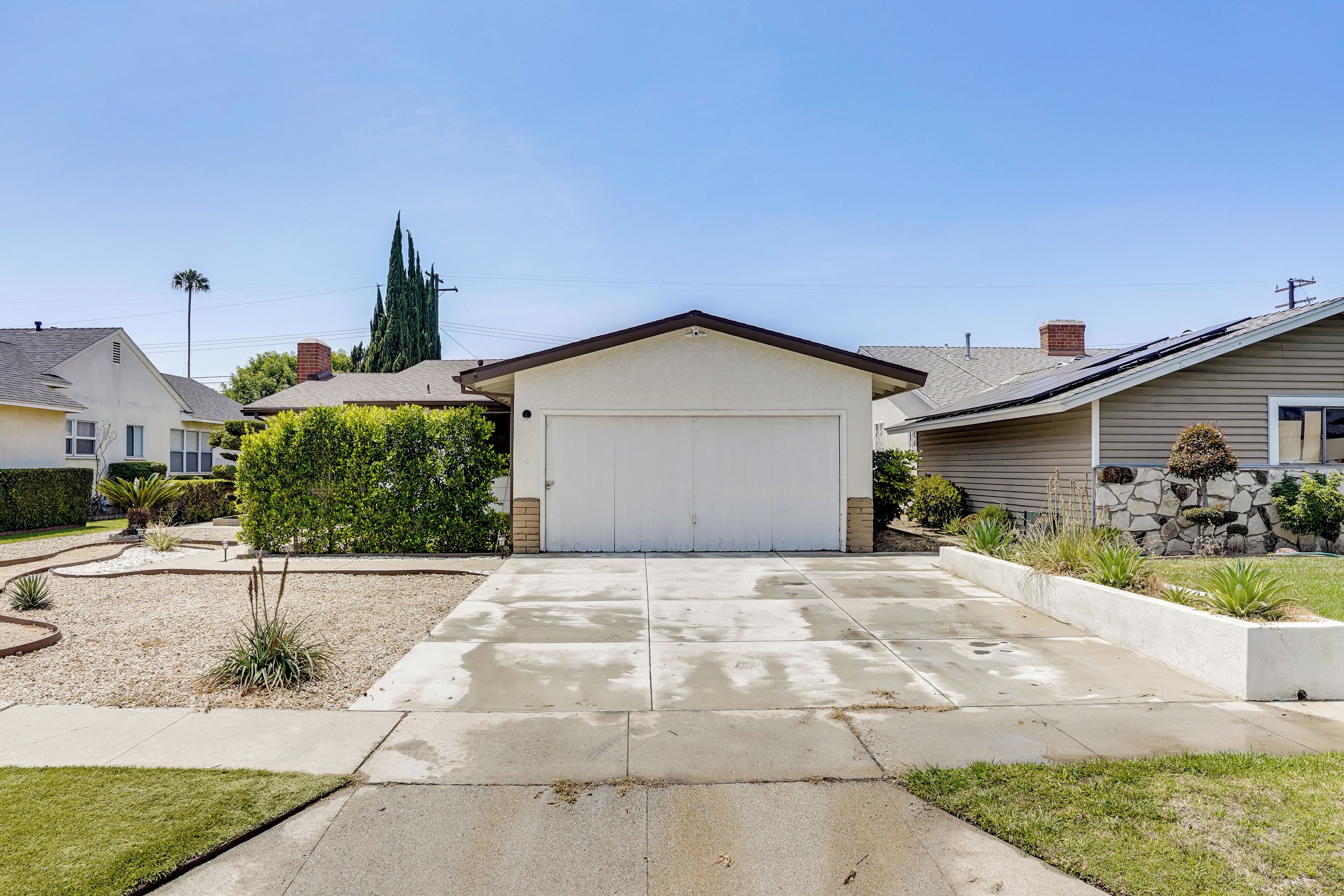 A residential driveway leading to a garage with a white garage door, surrounded by a landscaped garden with various plants and a small tree.