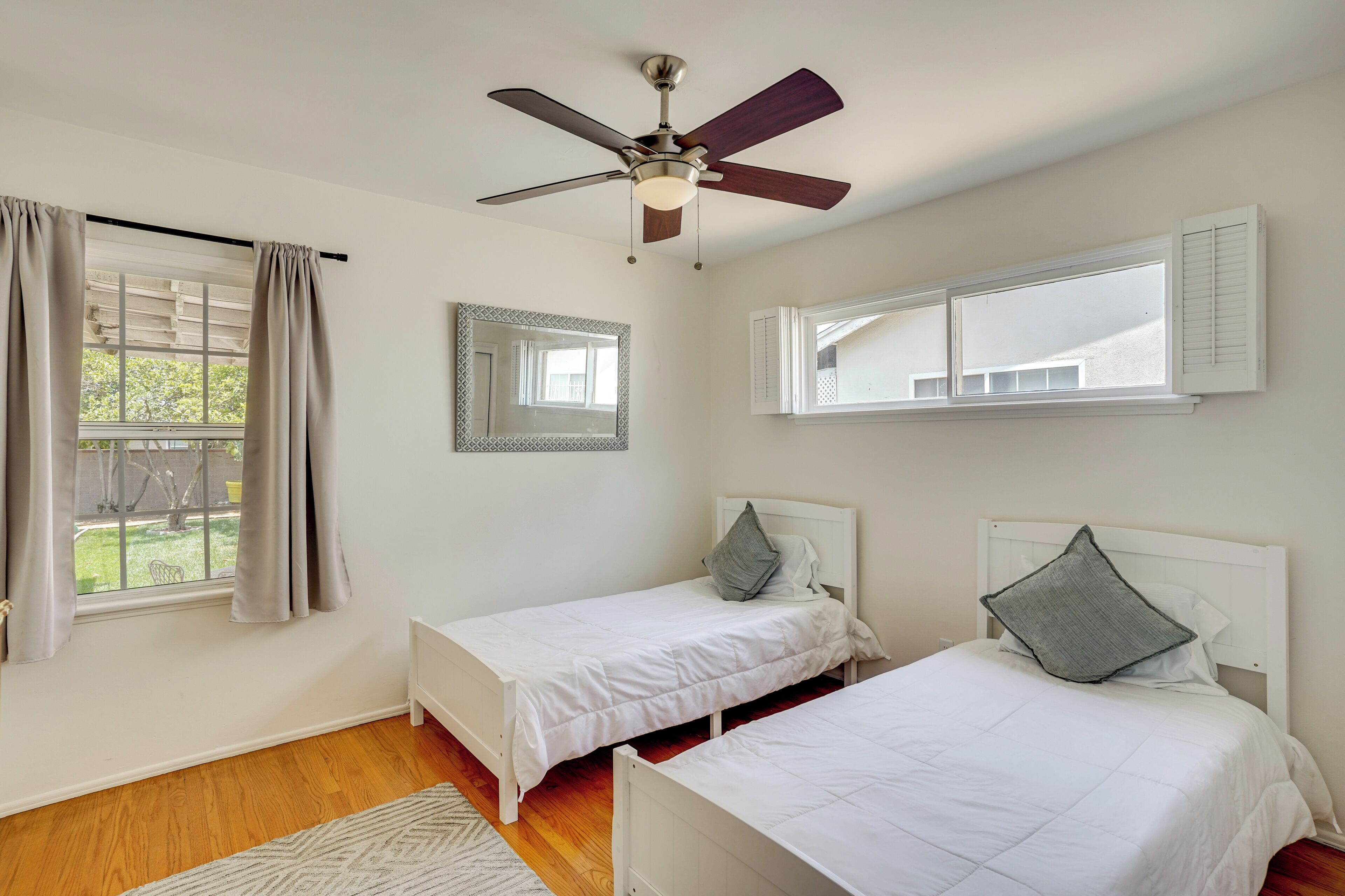 A bedroom with two single beds, a ceiling fan, a window with shutters, and a mirror on the wall.
