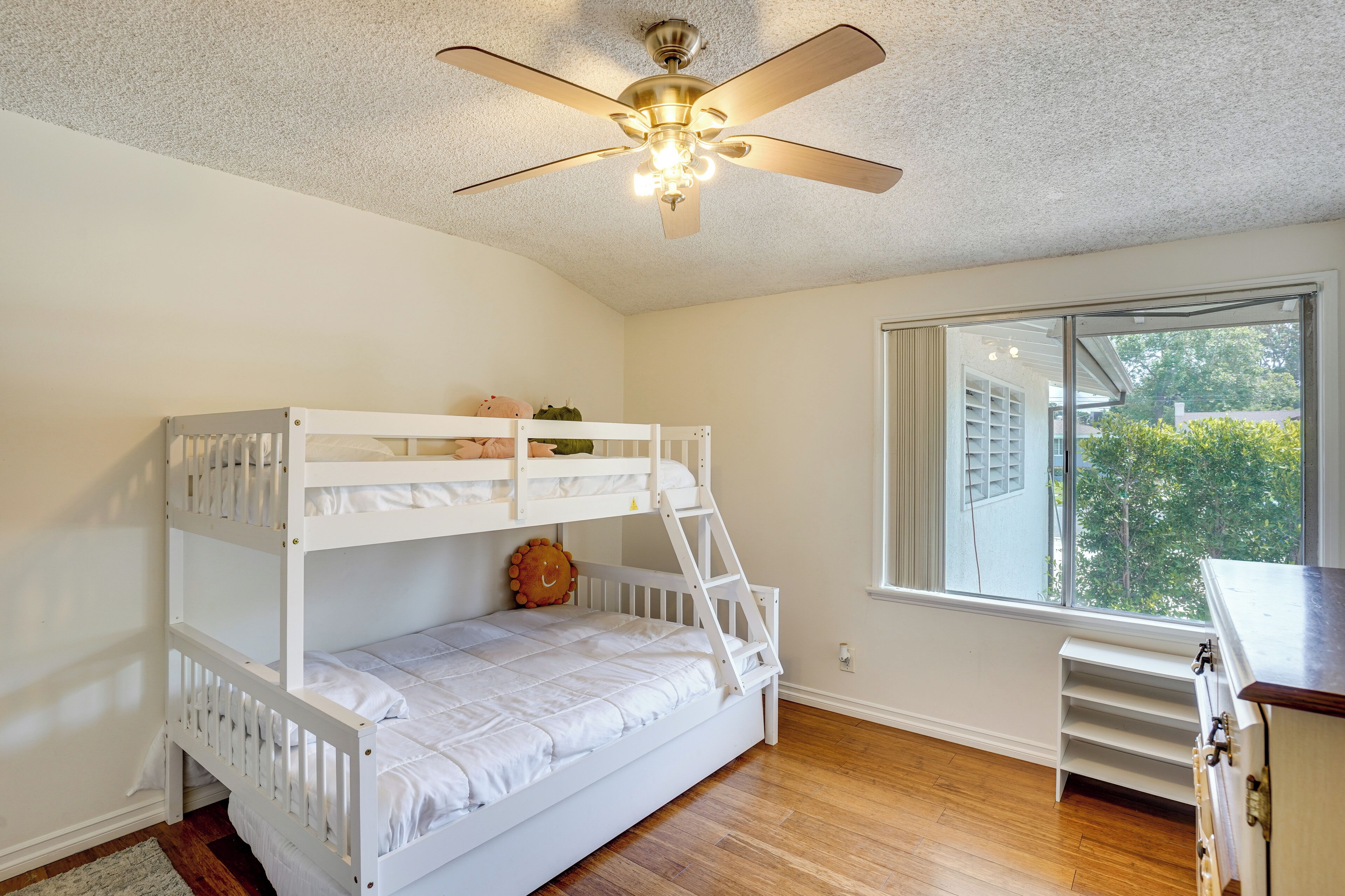 A bunk bed room with a ceiling fan, a window with blinds, and a wooden floor.