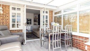 A sunlit room with a dining table and chairs, a sofa, and a brick wall. - Coastal Retreat With Hot Tub Walk to Beach (Chichester)