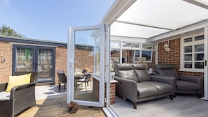 A covered patio with a glass door leading to a brick wall, a leather sofa, and a wicker chair. - Coastal Retreat With Hot Tub Walk to Beach (Chichester)