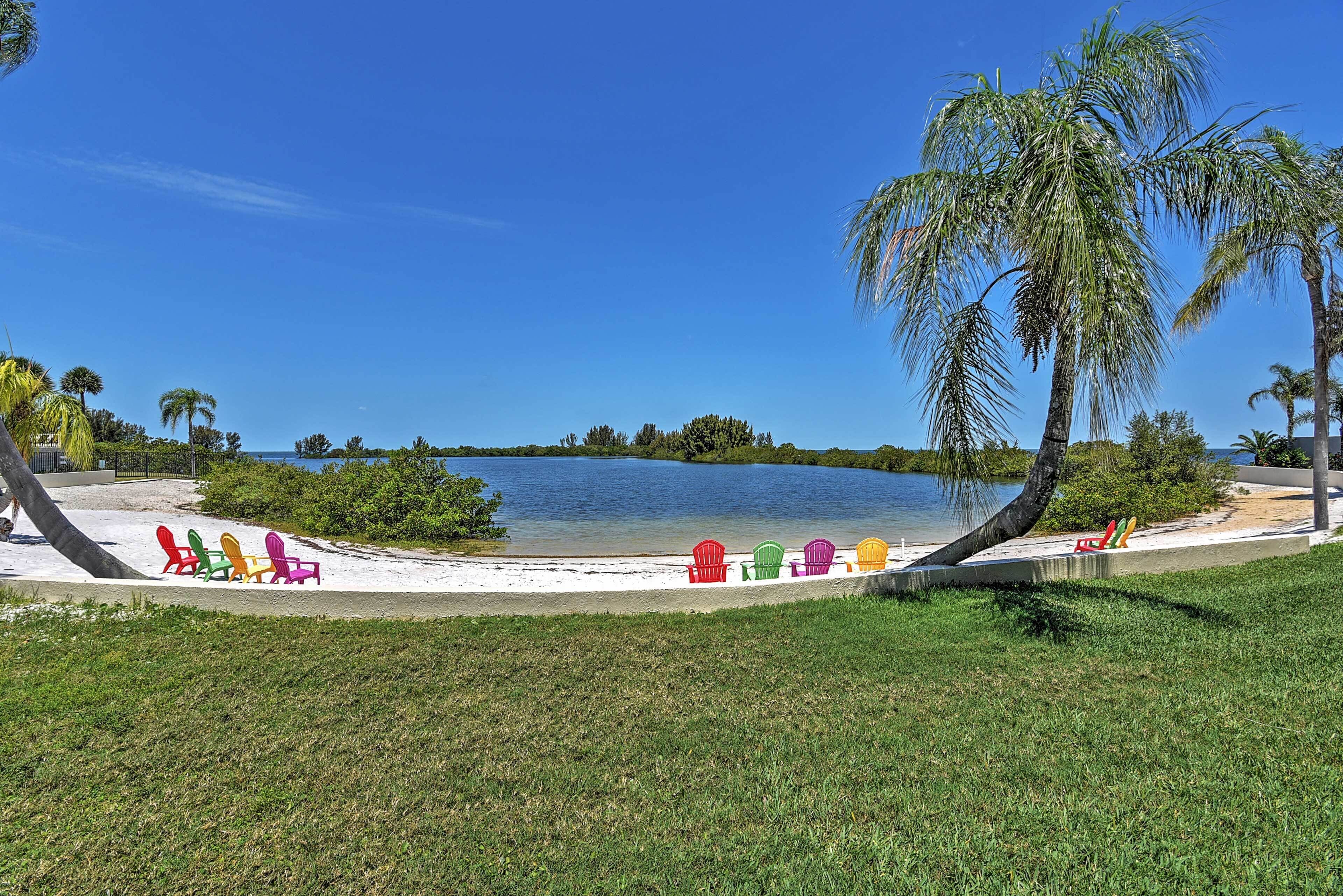 A beach with colorful Adirondack chairs lined up on the sand, a palm tree, and a calm lake in the background.