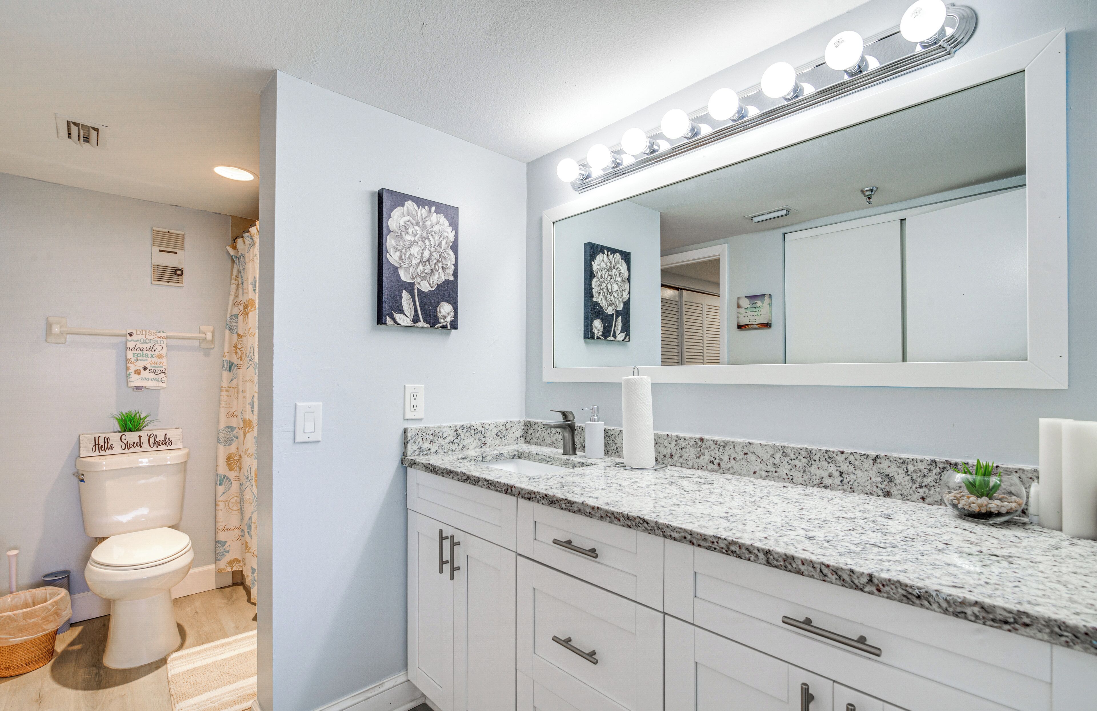 Un baño con inodoro blanco, lavamanos con encimera de granito y un espejo grande con luces superiores.