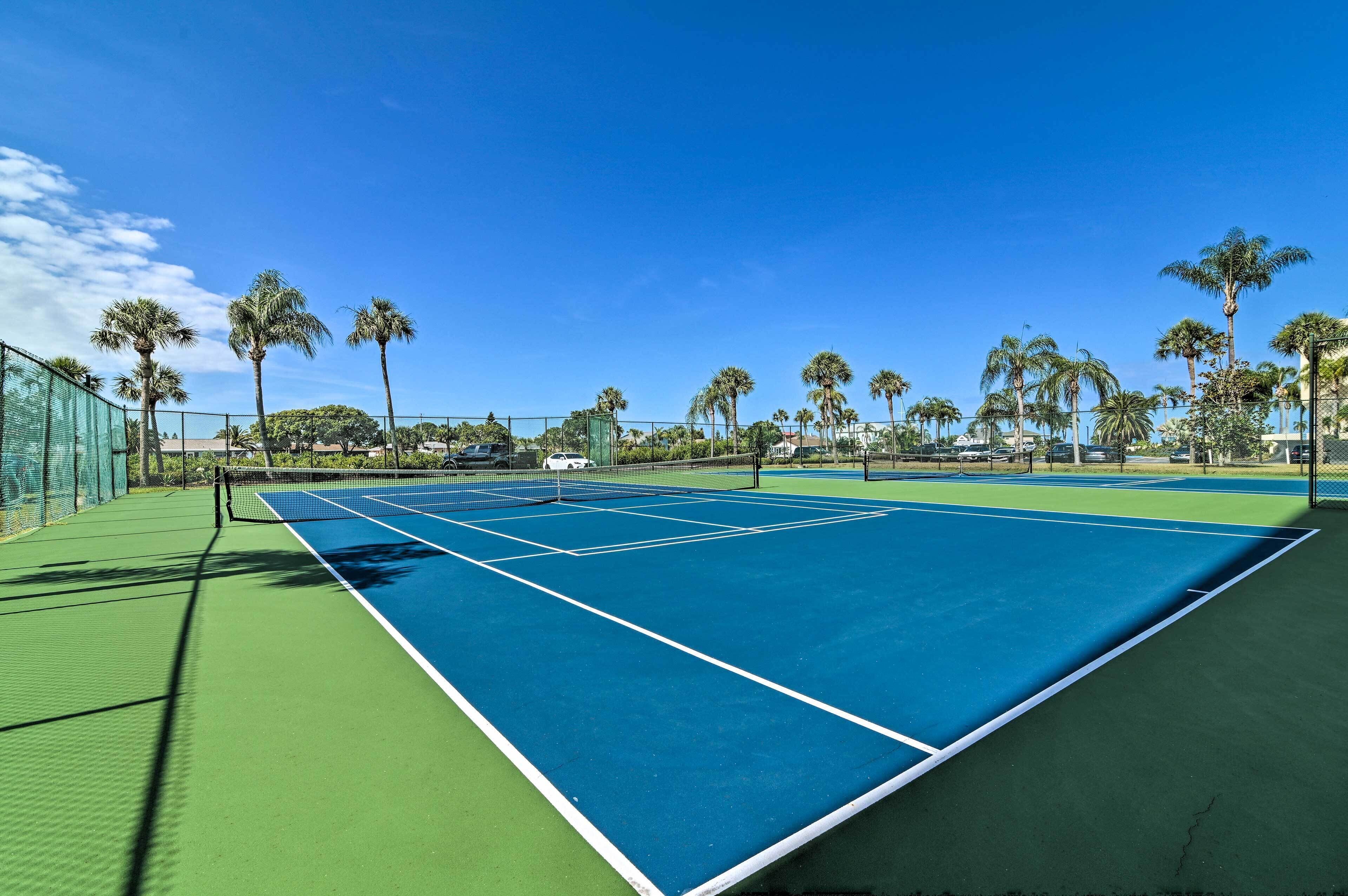 Un court de tennis avec une surface verte et bleue, entouré d’une clôture et de palmiers.