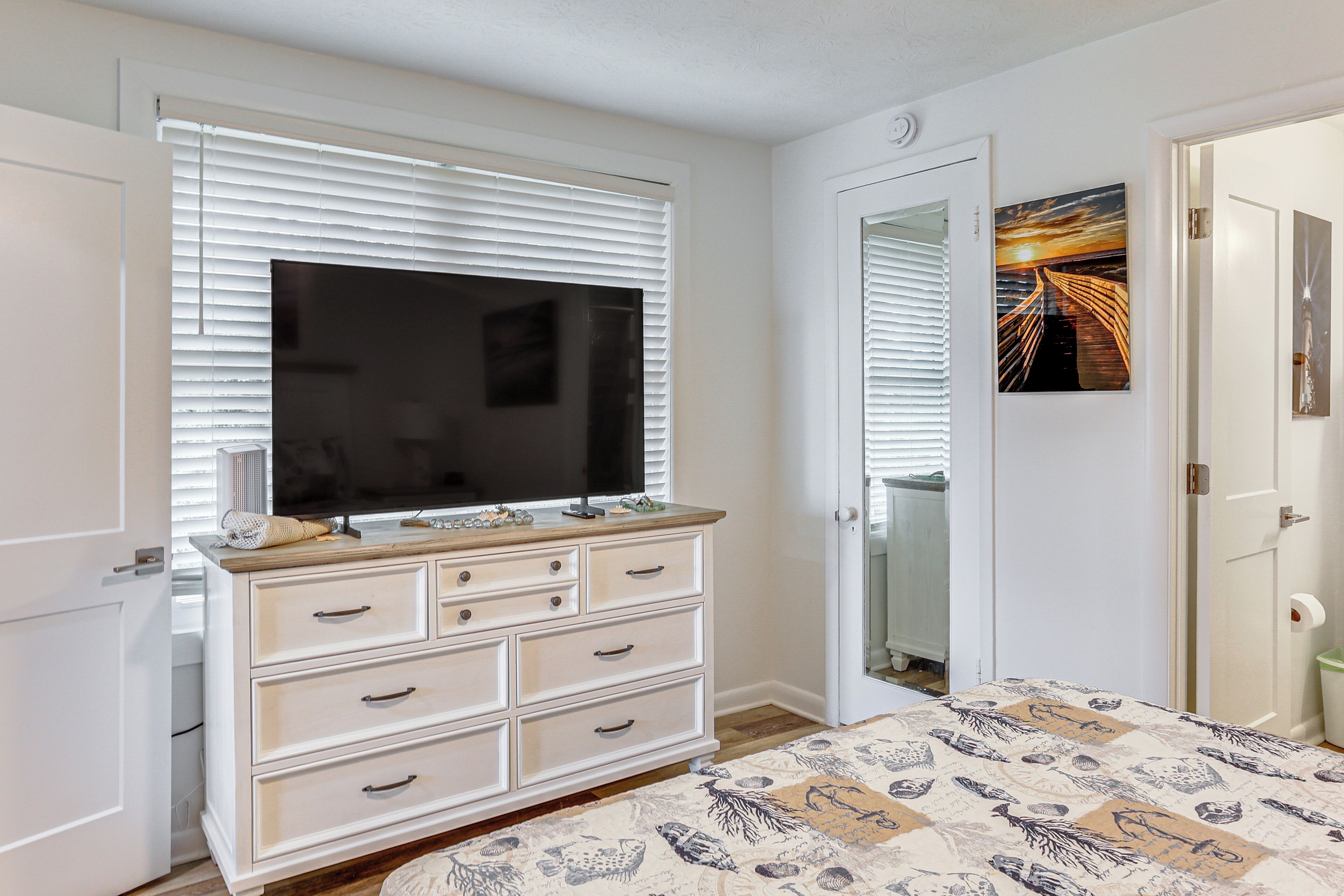 Une chambre à coucher avec une grande télévision posée sur une commode, un lit recouvert d’une couvre-lit à motifs et une porte donnant accès à une salle de bain.