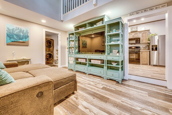 A spacious living room with a large beige sectional sofa, a teal storage unit, and a kitchen area visible through an open doorway. - Lakehouse With Sandy Beach and Boating (Belmont)