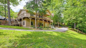 A two-story wooden cabin with a porch and a flag, surrounded by lush greenery and a paved driveway. - 5 Mi to Dtwn Hiawassee: Creek-front Home w/ Deck (Hiawassee)