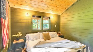 Ein Schlafzimmer mit einem Bett, zwei Fenstern, einem Nachttisch, einer Lampe und einer US-amerikanischen Flagge.