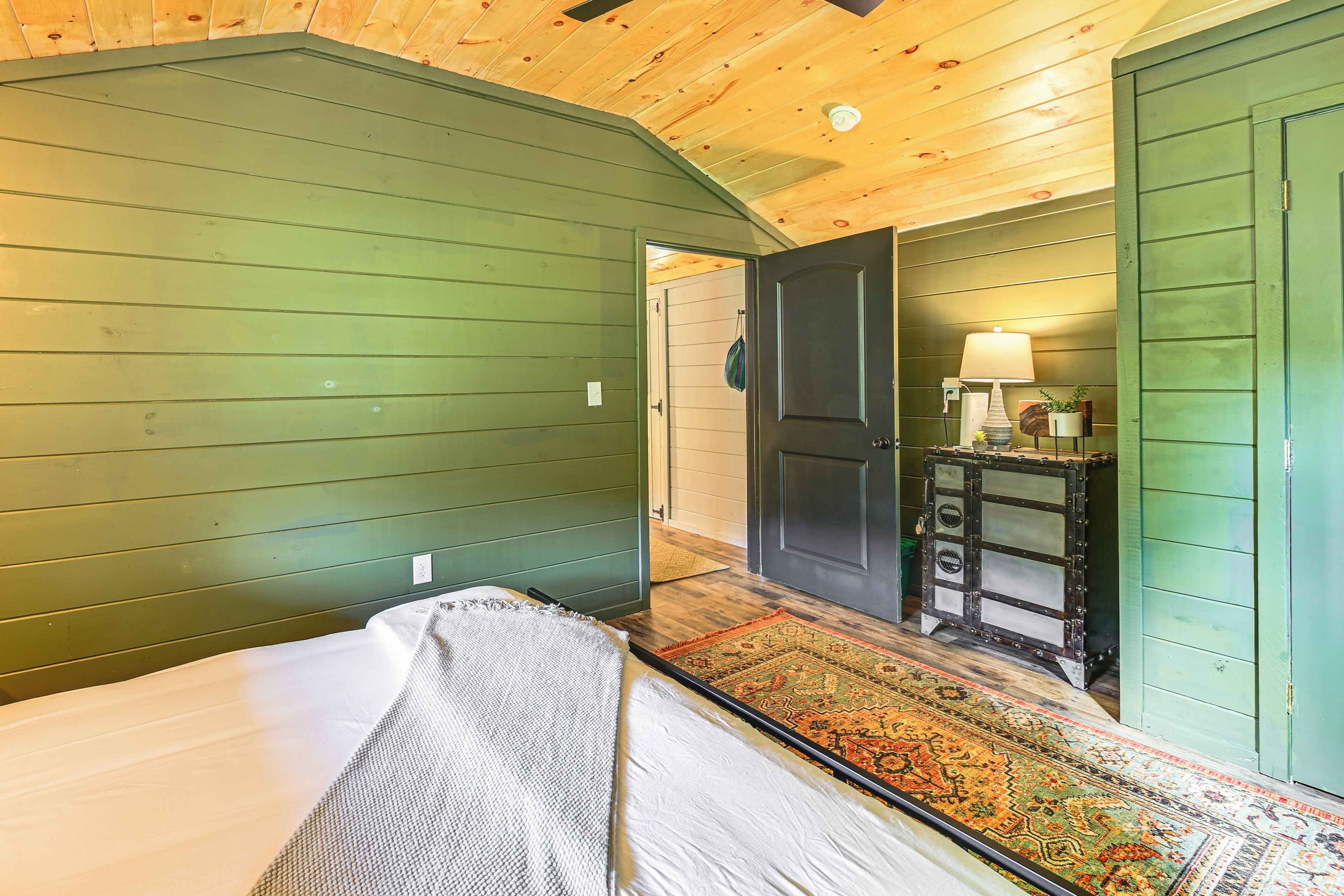 A room with a bed, a green wall, a wooden ceiling, a door, and a small table with a lamp.