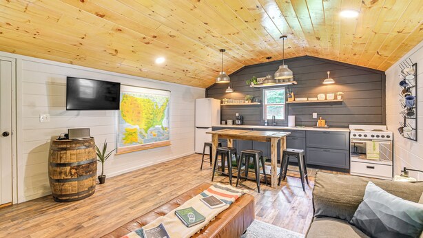 Ein gemütliches Wohnzimmer mit Holzboden, einem an der Wand befestigten Fernseher, einer großen Karte, einem Essbereich mit Hocker und einer Küche mit Herd.