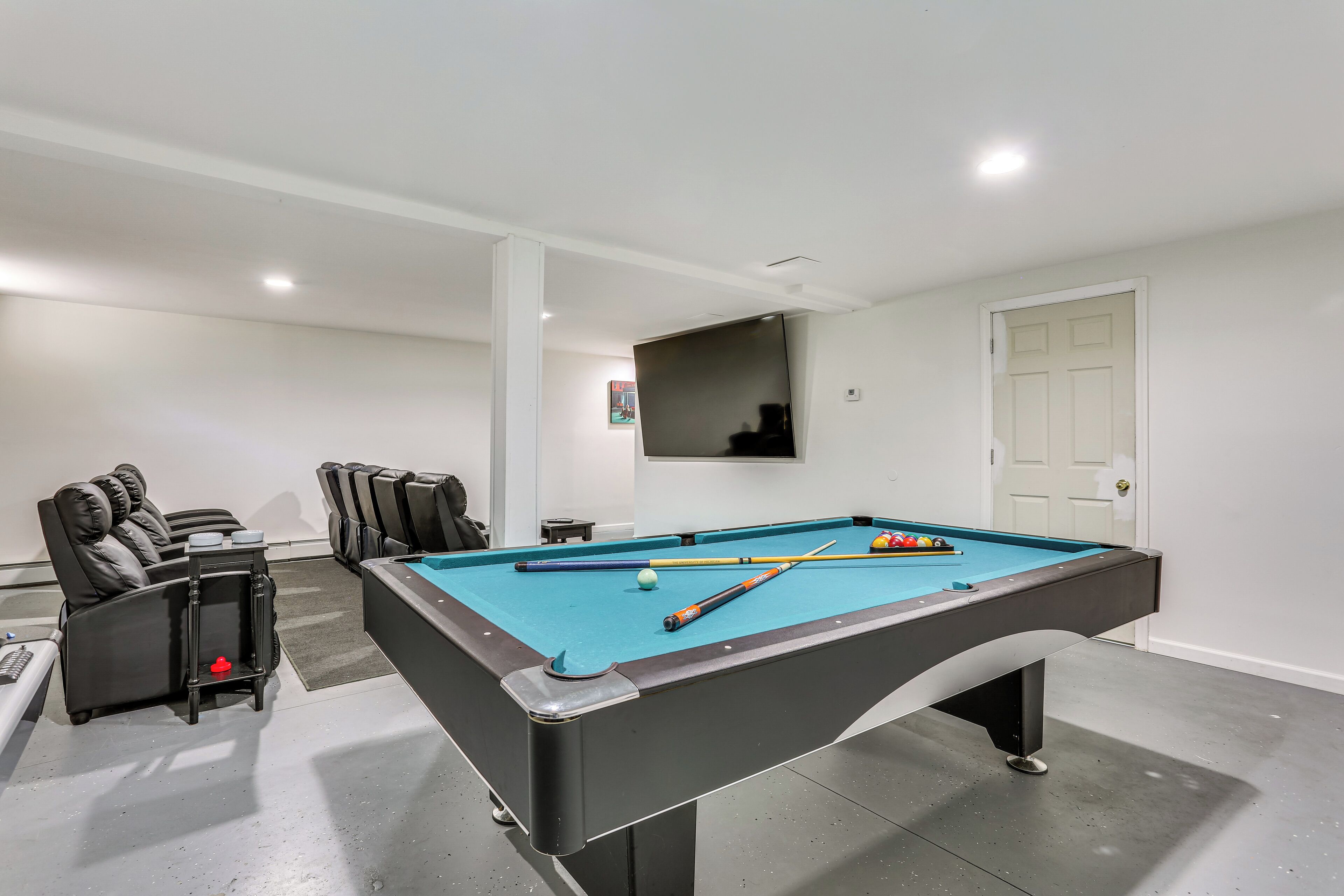 Une table de billard recouverte d’un drap bleu, entourée de fauteuils en cuir noir, d’une petite table et d’un téléviseur fixé au mur.