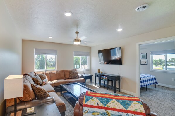 A living room with a sectional sofa, a coffee table, a TV mounted on the wall, and a window with blinds. - Missouri River Retreat < 1 Mi to the Water! (Pierre)
