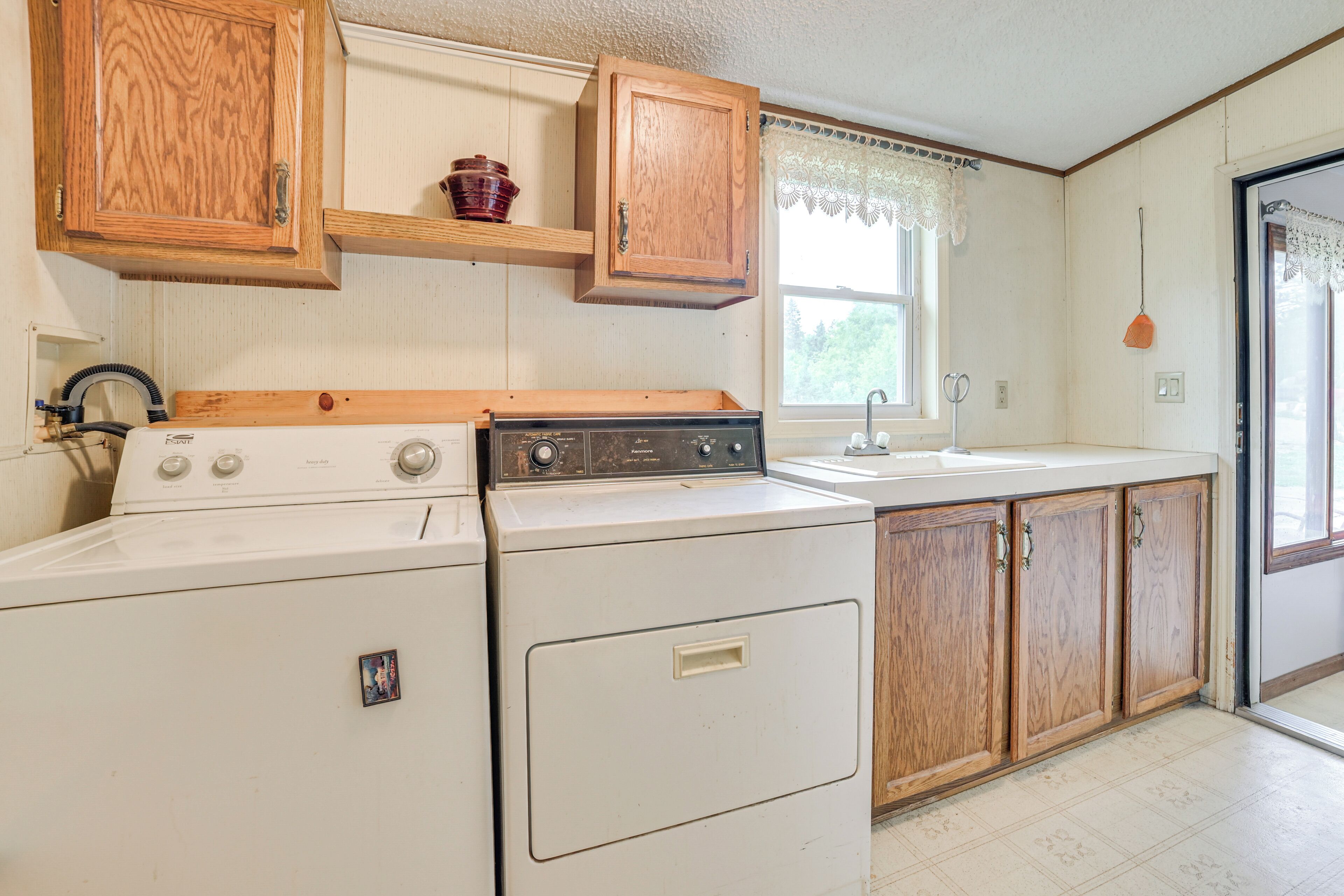 A laundry area with a washer and dryer, wooden cabinets, a window with lace curtains, and a sink.