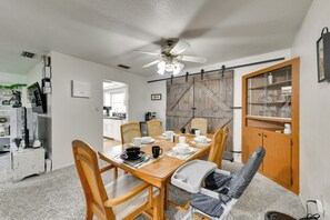 Un coin repas avec une table en bois, des chaises et un ventilateur de plafond. On aperçoit une cuisine au fond, avec une porte coulissante de style rustique.