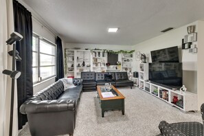 Un salon avec un canapé en cuir noir, une table basse en bois et un grand téléviseur à écran plat. On y trouve des étagères ornées de divers objets décoratifs et une fenêtre avec des rideaux noirs.