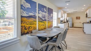 Un comedor con una mesa larga, sillas grises tapizadas y tres grandes cuadros de paisajes en la pared.