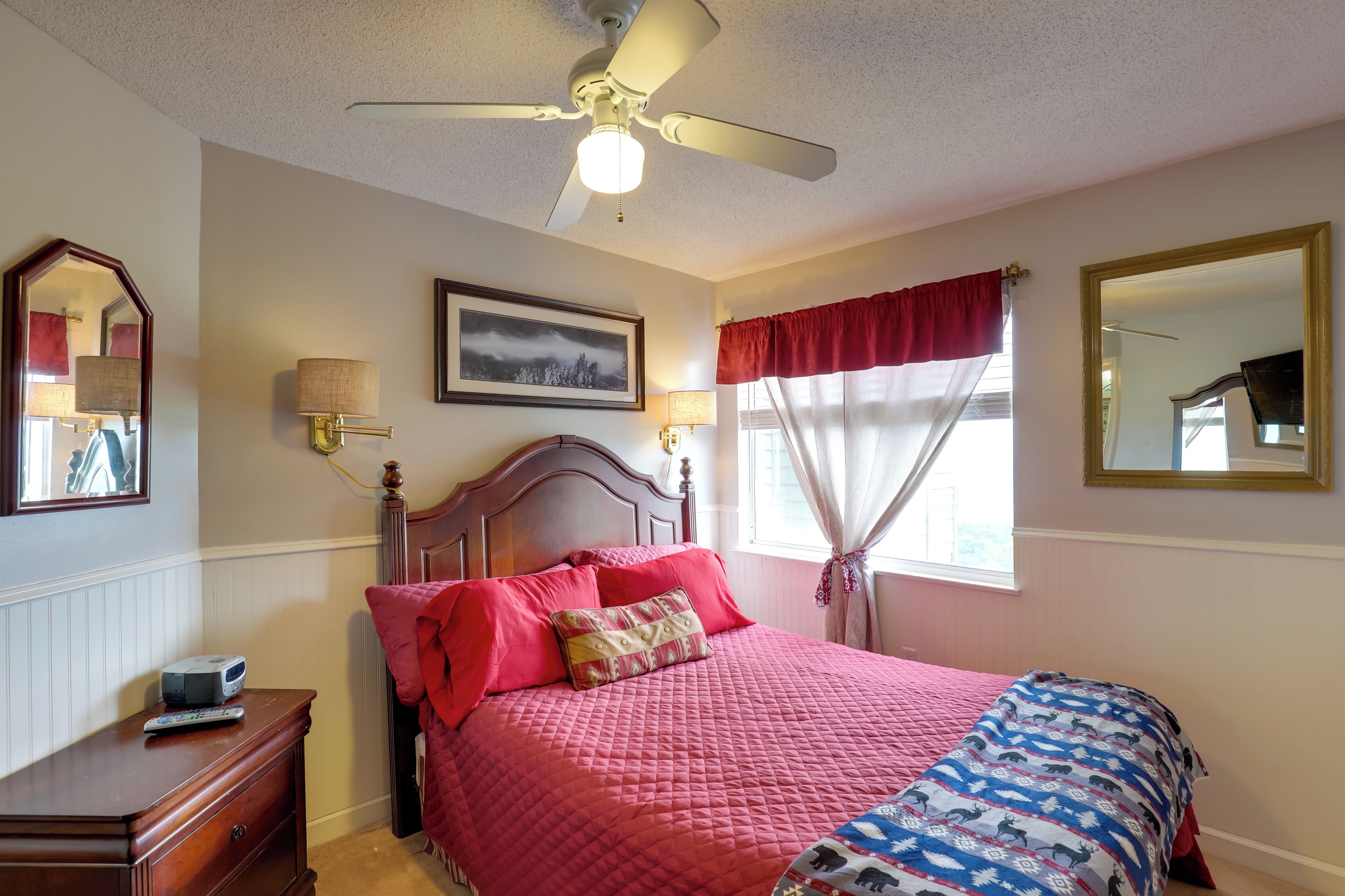A bedroom with a bed, a dresser, a ceiling fan, a window with curtains, and a wall-mounted lamp.