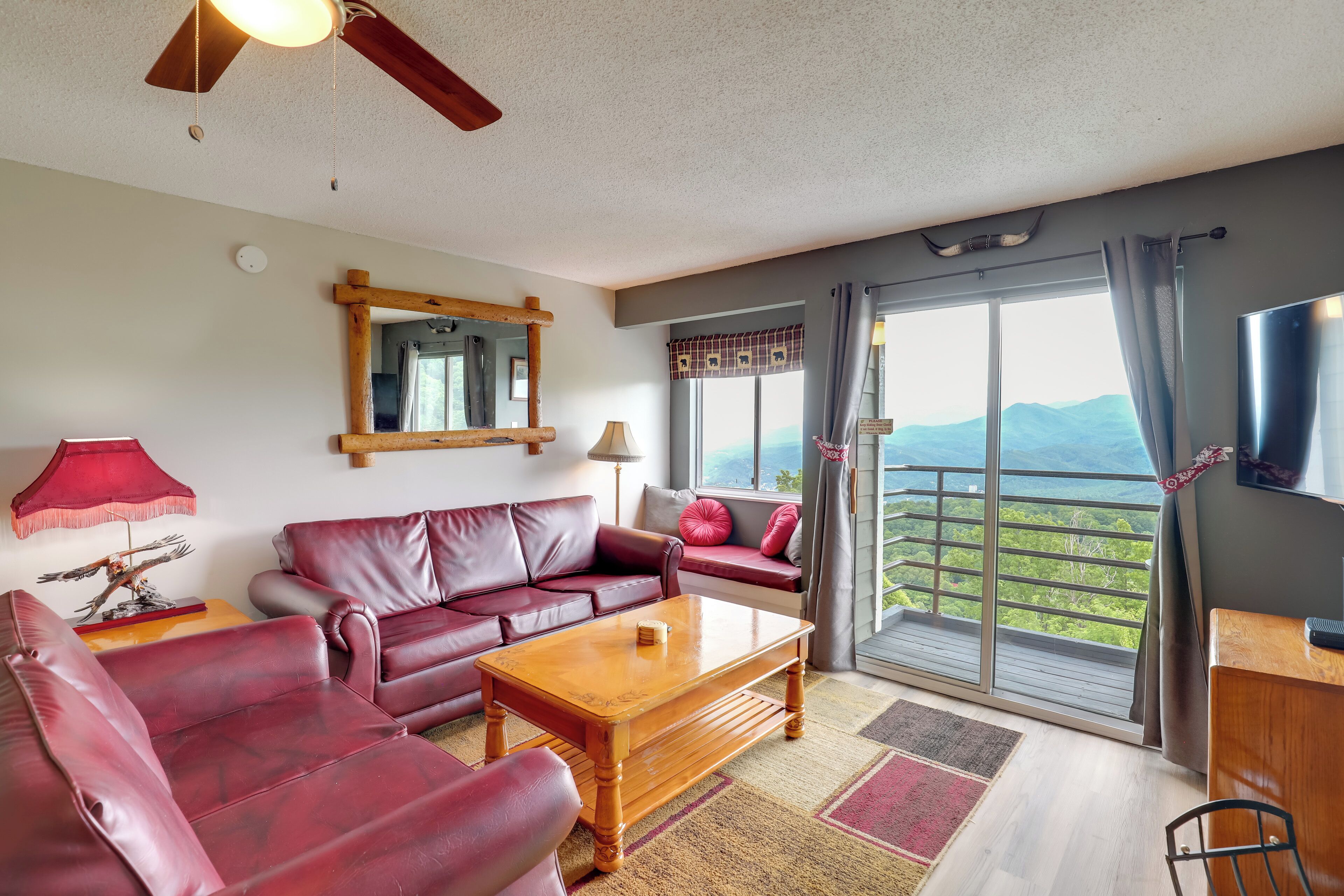 A living room with a red leather sofa, a wooden coffee table, and a TV on a wooden stand. There is a balcony with a view of the mountains.