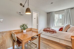 A room with a wooden dining table, a chair, a potted plant, a bed with a white mattress and pillows, and a window with curtains.