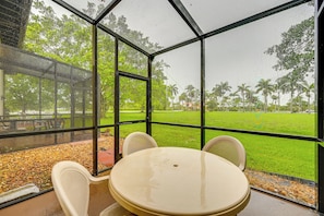 A glass-enclosed patio with a table and chairs, overlooking a green lawn and palm trees.