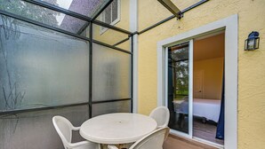 Une terrasse couverte avec une table et des chaises, une porte-fenêtre coulissante donnant sur une chambre, et un petit luminaire extérieur.