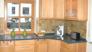 A kitchen with wooden cabinets, a window with a view of a fence and parked cars, a bowl of fruit, and a coffee maker. - Holiday Home 'Altmühltal Beilngries' with Mountain View, Private Terrace and Wi-Fi (Beilngries)