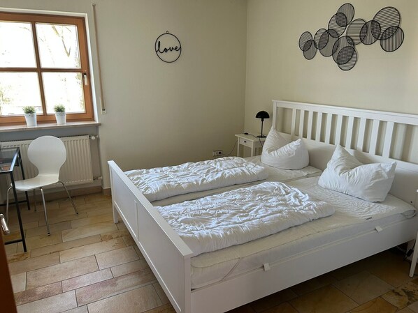 A bedroom with a double bed, a desk, a chair, a radiator, a wall decoration, and a window with plants.