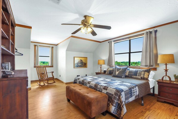 A bedroom with a large bed, a ceiling fan, a rocking chair, a wooden dresser, and a window with a view of greenery.