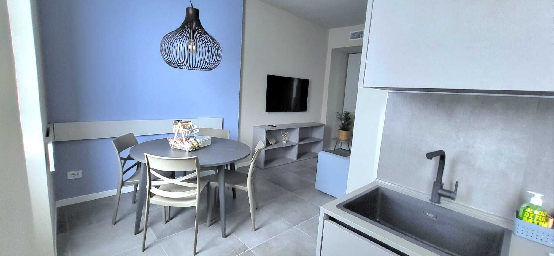 Eine kompakte Küche mit integriertem Spülbecken, ein Essbereich mit rundem Tisch und Stühlen sowie ein an der Wand montierter Fernseher.